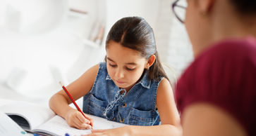 A young girl writes in her notebook, and her mother watches her, showing encouragement and support.