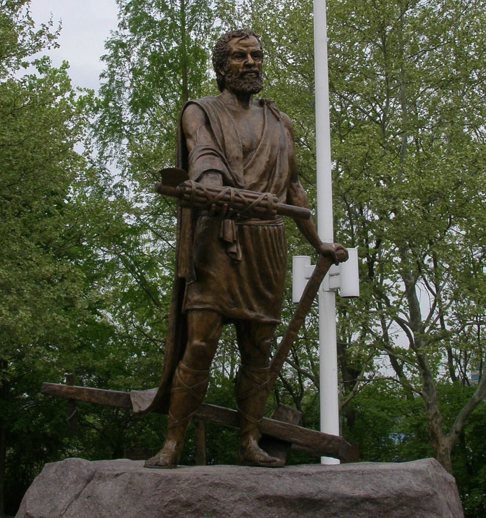 Bronze statue of a bearded man holding a fasces, set on a rocky pedestal. Surrounded by trees, it conveys a solemn, historic mood.