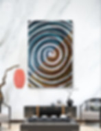 A gray, blue, orange, and black swirly painting. The parts that swirly in blue and black and the part that swirls in black and orange are split triangularly. The bottom triangle has gray, black, and orange. The top one has gray, black, and blue. It is set in a home setting in front of a white wall, with a gray coffee table, a predominately black table with drawers and a plant on top, and a light gray chair on the side.