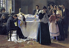 Mourners gather around a flower-adorned casket in a dim room. A man in black slumps in grief as others stand with somber expressions.