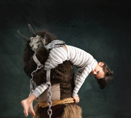 Child in striped pajamas carried by costumed figure with a horned mask. Dark background creates a mysterious, whimsical mood.