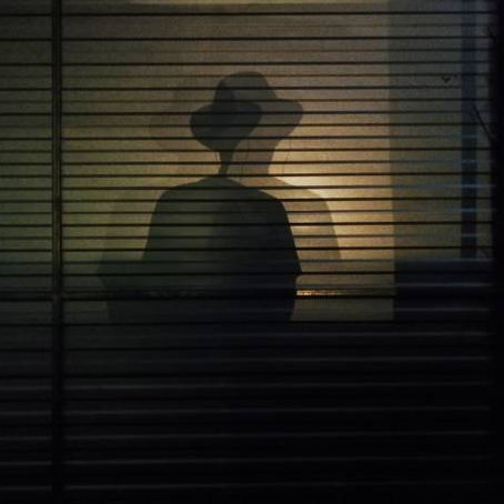 Shadow of a person wearing a hat cast on a window with blinds, dimly lit from behind, creating a mysterious and moody atmosphere.