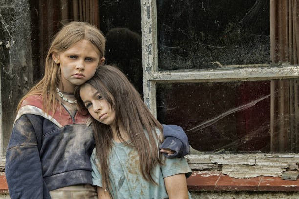 Two girls with somber expressions stand in front of an old, cracked window. One has an arm around the other. Their clothes are worn.