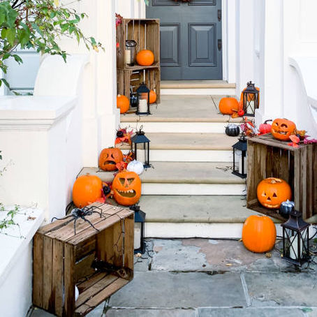 Pumpkins with carved faces and lanterns decorate white house steps. Wooden crates hold more pumpkins. Autumn ambiance, no visible text.