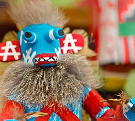 Blue Kachina doll with red arms and feathered collar, featuring bold patterns. Vibrant, colorful background creates a lively atmosphere.
