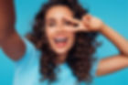 Young brunette woman with curly hair smiling big and holding a hotizontal peace sign in front of her left eye