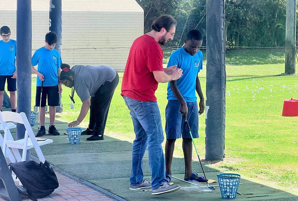 A man in a red shirt teaches a boy in a blue shirt to golf at Keiser University's driving range. Other students practice nearby. Sunny day, green grass.