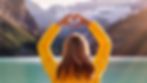 Woman in a yellow jacket holding heart hands over her head with a lake and mountains in the background