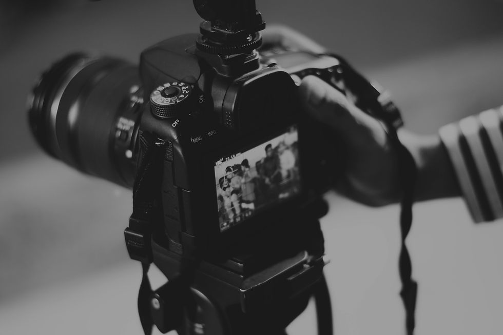 Close-up of a camera screen showing a blurred group photo. A hand holds the black camera with a striped sleeve visible. Monochrome image.
