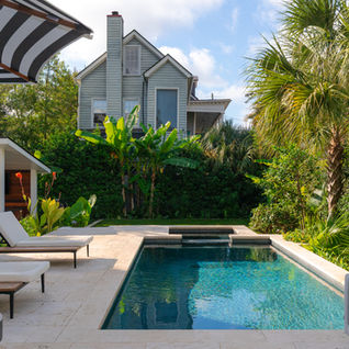 An wide view of a downtown Charleston backyard landscape with a rectangle pool. The pool is flanked by a white patio, turf, and lush landscaping. The left corner shows a peek of the outdoor kitchen with warm wood tones and sleek lines. Tropical plants like fan plams, shell ginger, and canna lilies install by Meeting Green.