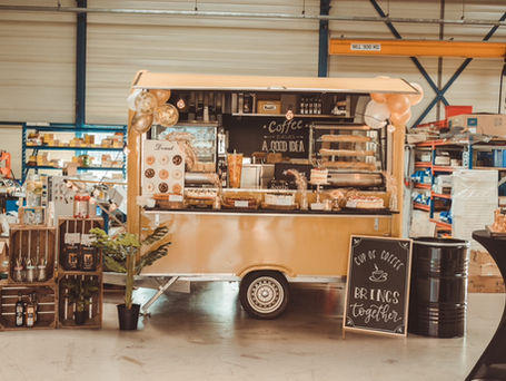 Gelber Foodtruck mit Donuts und Getränken in industrieller Umgebung. Schilder mit "Coffee", Luftballons und Pflanzen dekorieren den Stand.