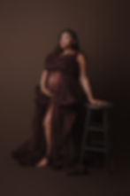Pregnant woman in a flowing, dark brown gown leans on a stool, set against a matching brown background after getting a prenatal massage in montgomery al