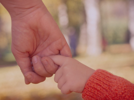 An adult and child hold hands against a blurred background.  Child wears orange.