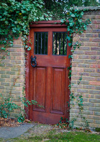 Rustic red brick walls frame a dark brown wooden gate, partially covered in ivy, with a wrought iron lion designed door handle and railings to peer through.