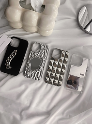 Four stylish and unique iPhone 14 cases displayed on a white surface. From left to right: a black case with a chunky silver chain, a silver case with a textured, cracked mirror design, a Silver case with a geometric chocolate like pattern, and a clear case with a playful cat photo.