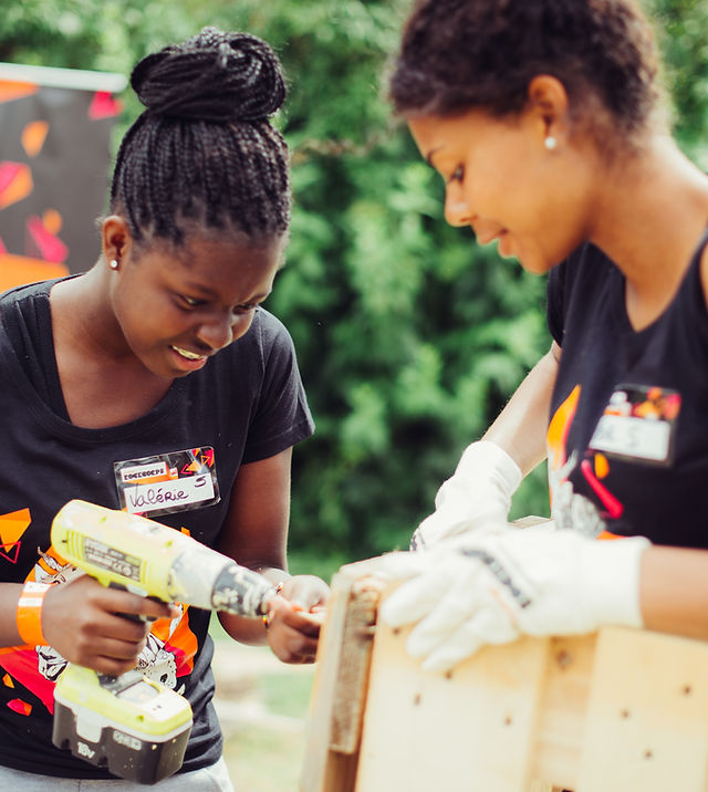 Two black teenager girls volunteering - one is holding a drill, the other the wood. Both wearing RockCorps t-shirts.
