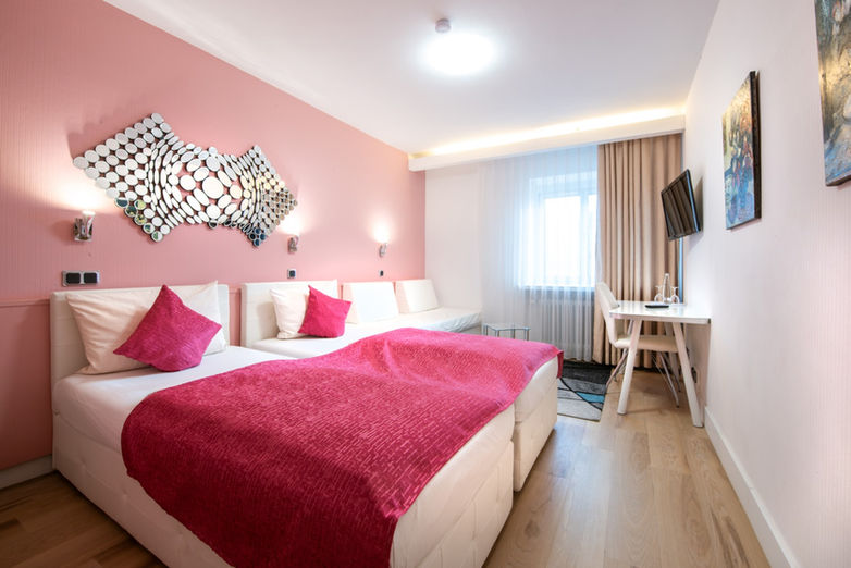 Modernes Zweibettzimmer mit pinkfarbener Wand, zwei weißen Betten, pinkem Bettüberwurf und Schreibtisch am Fenster.