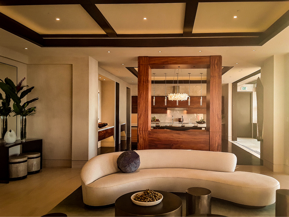 Image shows a stylish living area with a contemporary design, featuring a curved, cream-colored sofa and a set of modern, round coffee tables. The space is highlighted by warm wooden accents, a coffered ceiling with recessed lighting, and a sophisticated kitchen in the background with a central island and elegant pendant lights.