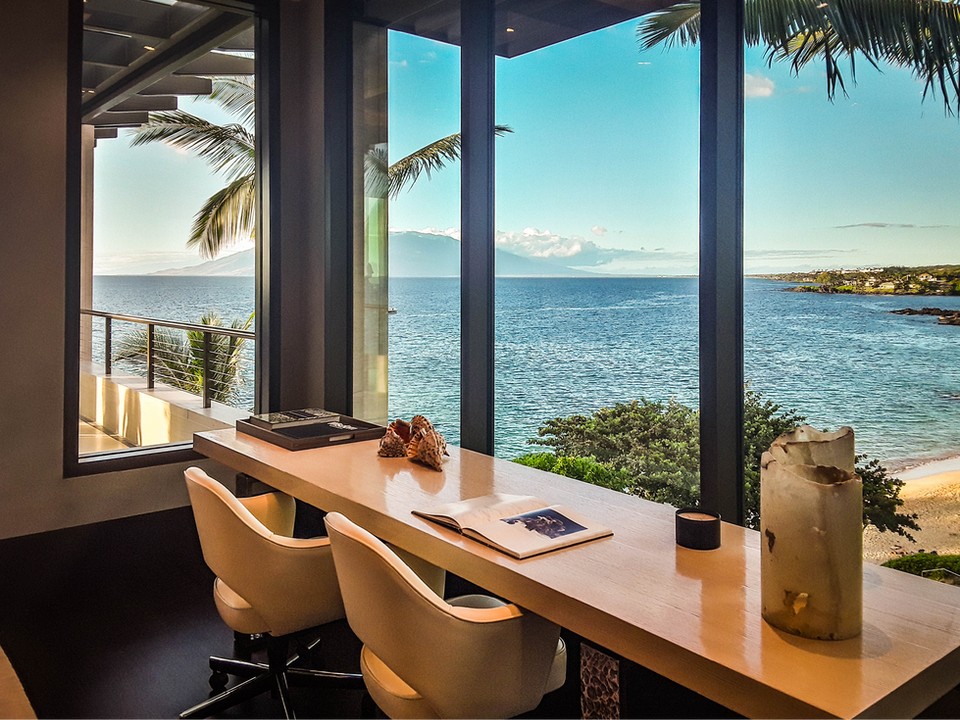 Image displays a serene workspace with a sleek desk and two chairs, positioned by large windows that offer a breathtaking view of the ocean, palm trees, and the coastline.