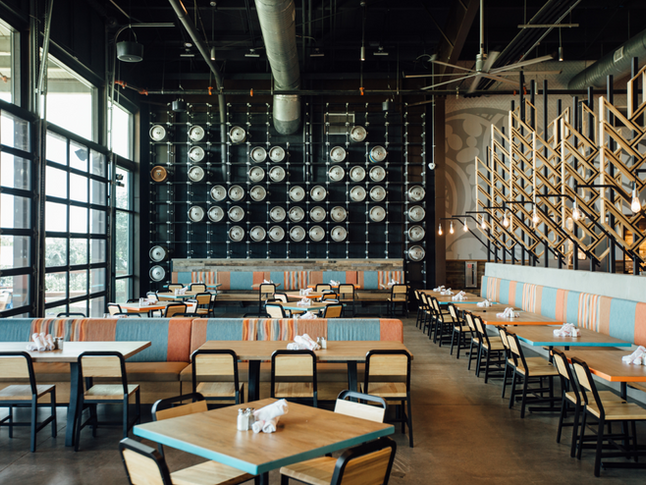 A brewery dining area with cane-back chairs, teal and orange upholstered booths, a dramatic wall of stacked barrel ends, and large industrial windows.