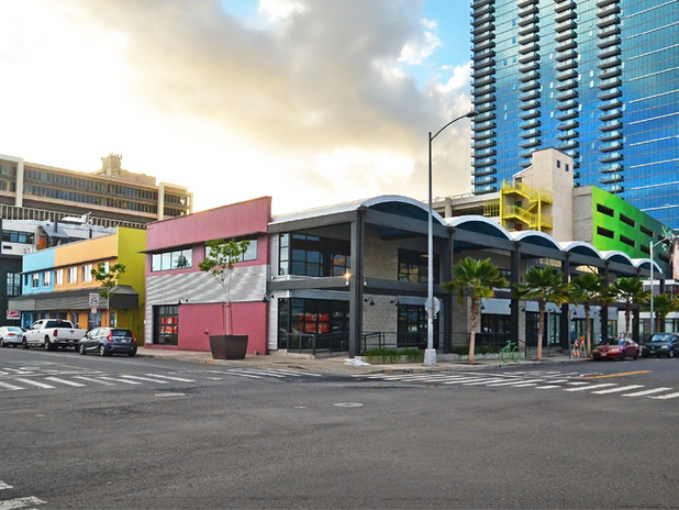A wide view of the SALT complex in Kakaʻako, highlighting colorful low-rise commercial buildings with a modern high-rise in the background. The architecture blends industrial and contemporary elements.