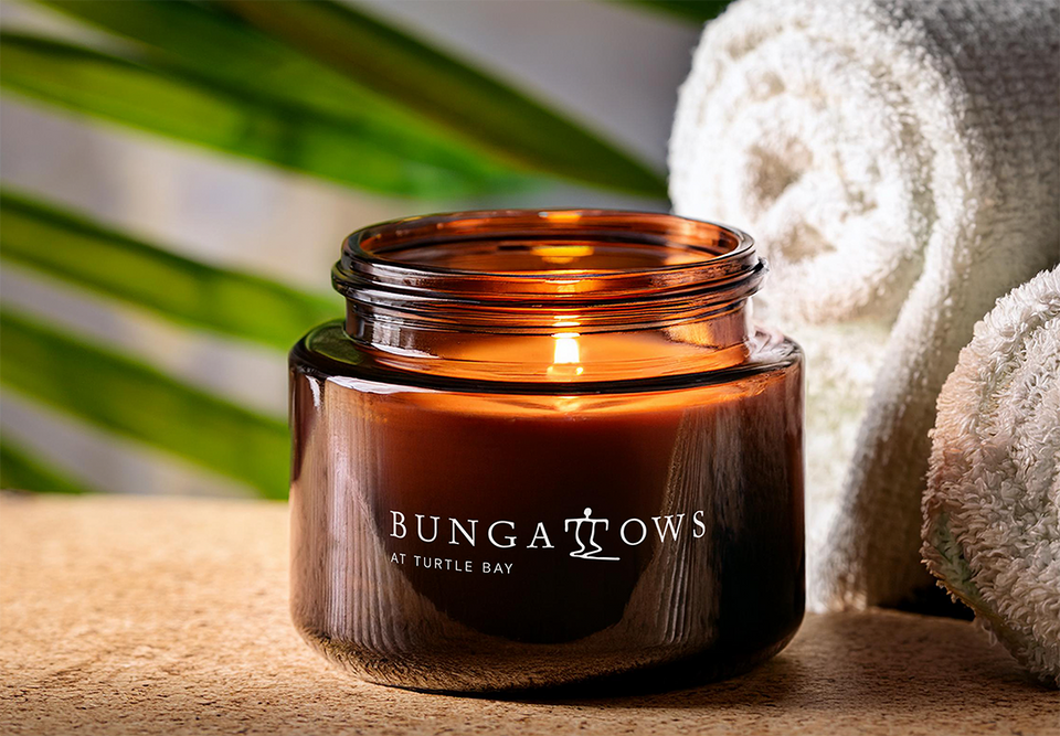 A Bungalows Turtle Bay branded amber glass candle jar with tropical greenery and rolled white towels in the background.