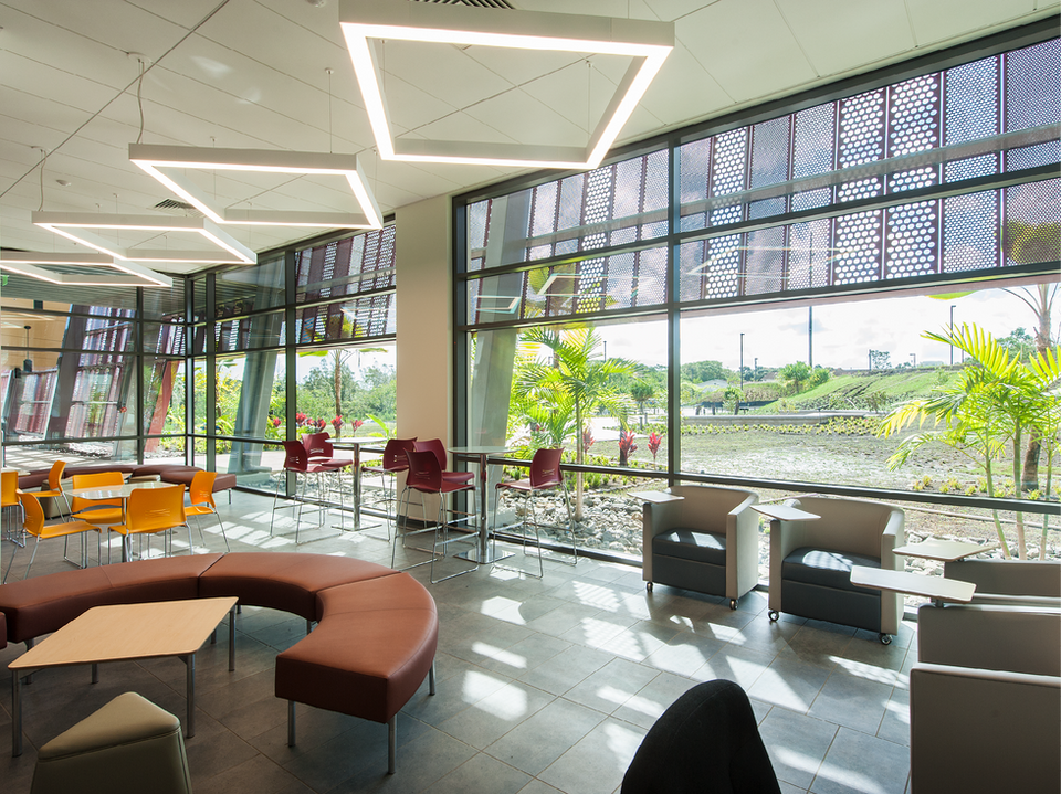 A bright school student lounge or common area with curved upholstered seating, geometric ceiling light fixtures, perforated screen walls, and large windows with garden views.
