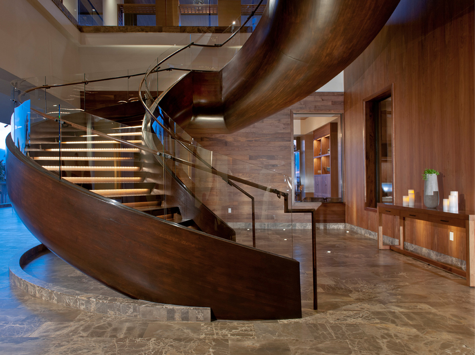 A grand, curved wooden staircase in a hotel lobby, featuring glass railings and ambient lighting. The sleek design harmonizes with the marble flooring and wooden wall paneling.
