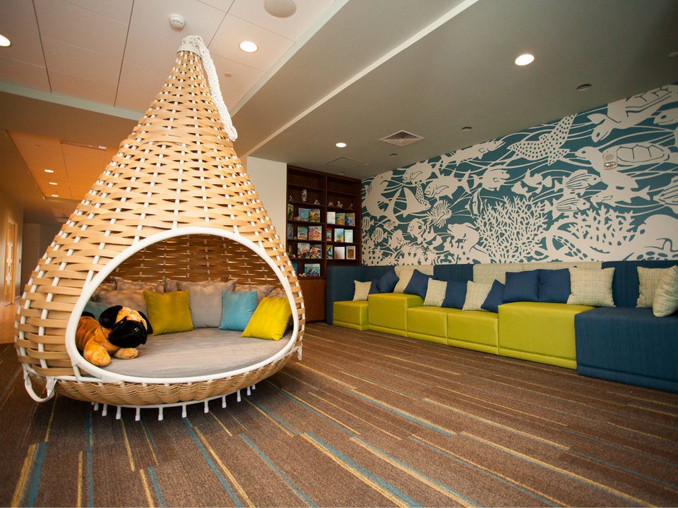 Image features a whimsical indoor play area with a large, teardrop-shaped wicker seating pod, colorful cushions, and a mural depicting underwater scenes.





