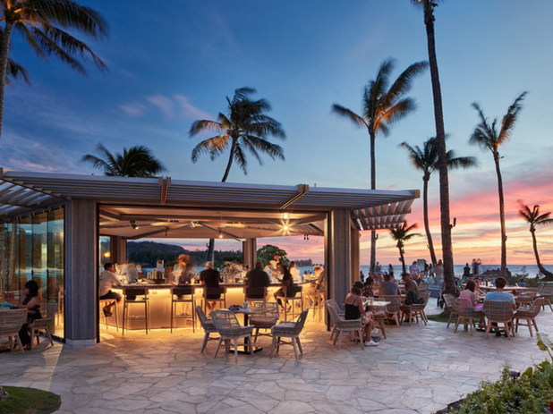 A lively outdoor bar and dining area by the beach at sunset, with patrons enjoying the vibrant atmosphere under palm trees