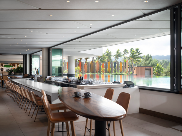 An open-air bar with modern seating overlooking an infinity pool and surfboard art installation, set against a tropical backdrop.
