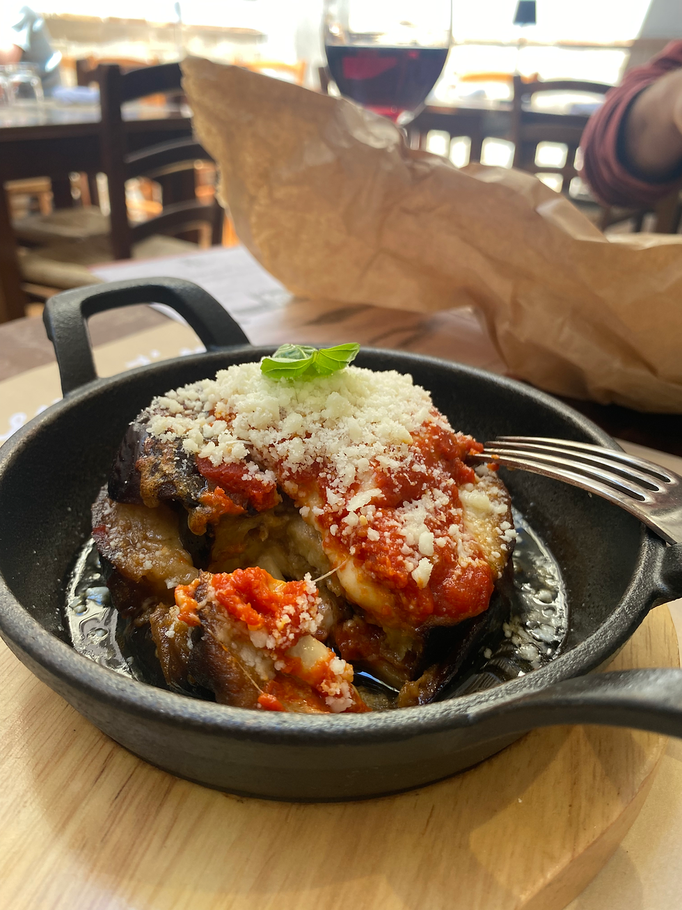Eggplant parmesan in a black skillet on a wooden table, topped with tomato sauce and cheese, with a fork and a glass of red wine nearby.