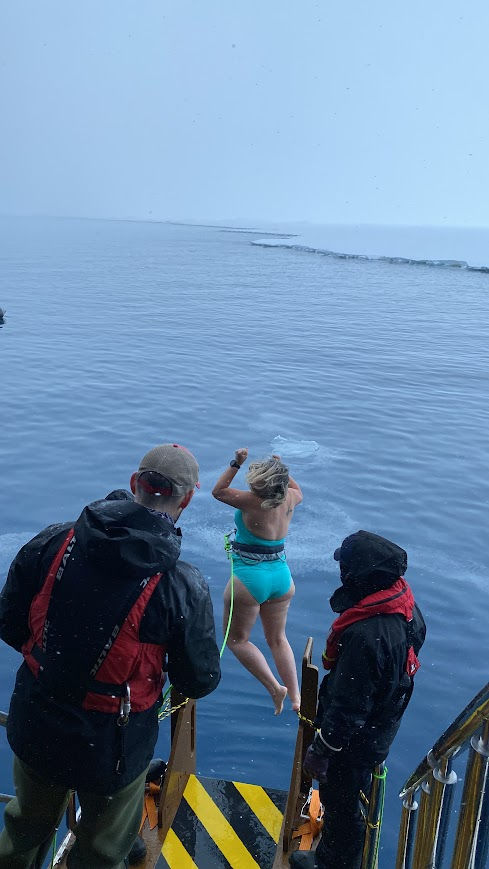 Person in turquoise swimsuit dives into the icy Antartic sea from boat, two others in winter gear watch. Overcast sky, safety ropes visible. Adventure mood. Welcome to the Polar Plunge!