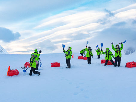 People in bright green jackets wave blue shovels on a snowy landscape with red bags. Overcast sky and mountains in background. Energetic mood.