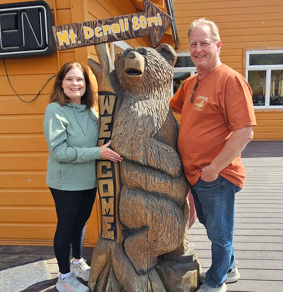 Travel advisor Ann and her husband smile beside a carved wooden bear holding a Denali sign.