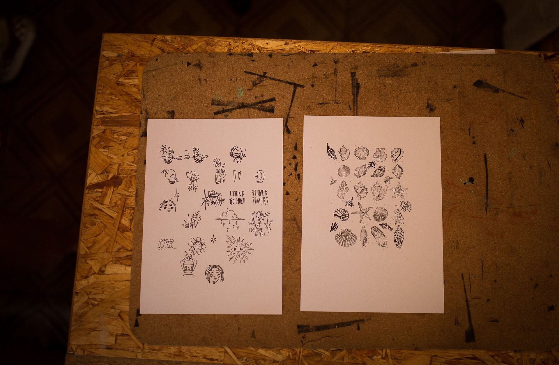 Two sheets of flash tattoo designs laid out on a wooden surface, showcasing various black ink illustrations, including flowers, shells, stars, and other nature-inspired motifs.