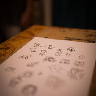 Close-up of a sheet of flash tattoo designs in focus, with the camera angled slightly to give a depth-of-field effect, showing nature-inspired doodles and text in black ink.