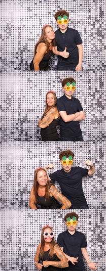 A brightly lit display of South Asian and British wedding guests interacting with a touchscreen DSLR selfie photobooth set up with a vibrant modern backdrop, holding funny masks while multiple photos loop on screen, alongside a demonstration of instant digital sharing and custom-designed 2x6 printed strips.