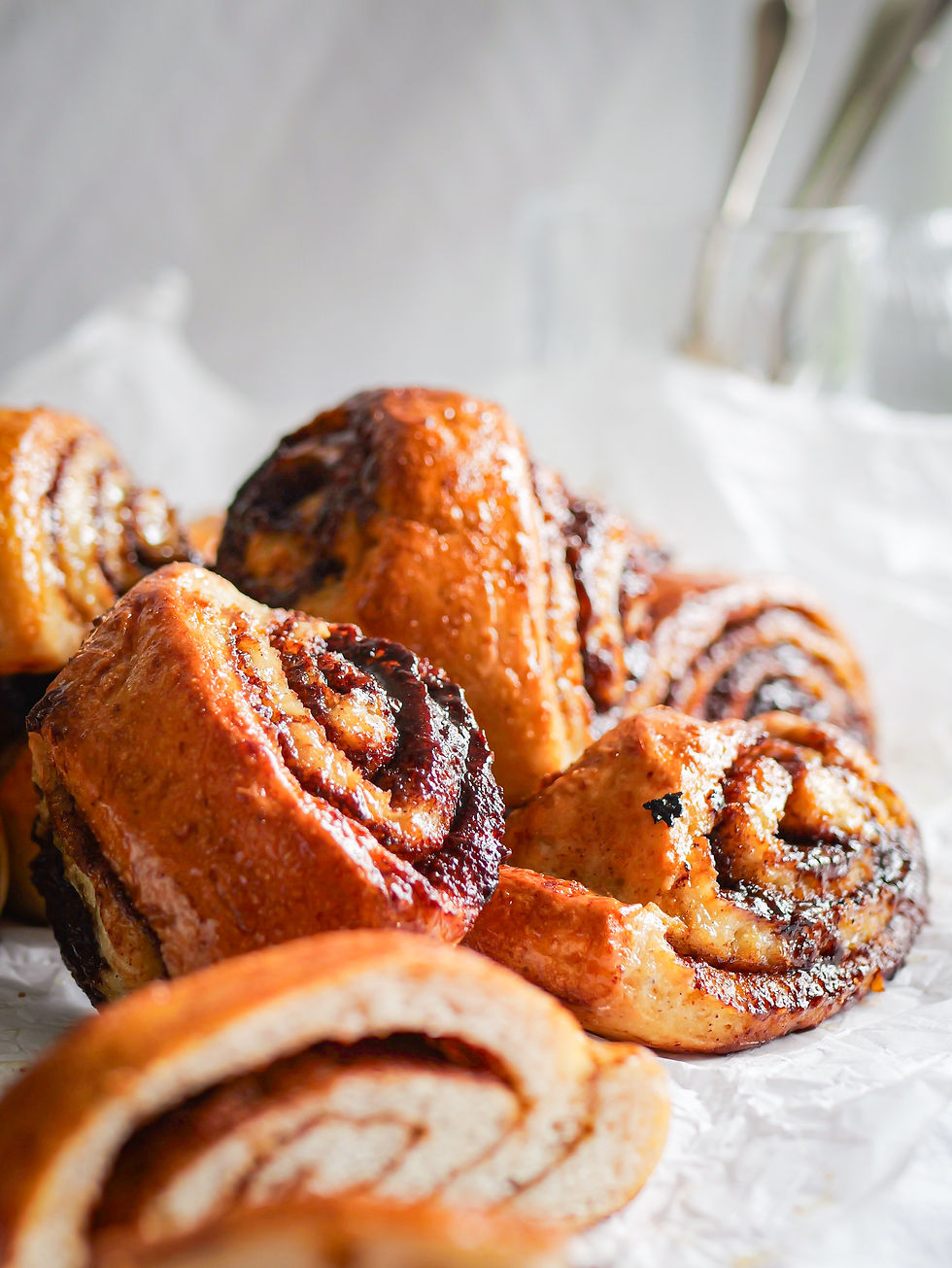 A pile of Gluten Free Finnish Cinnamon Buns (Korvapuusti) in front of a white background. Some glassware and cutlery is visible in the background.