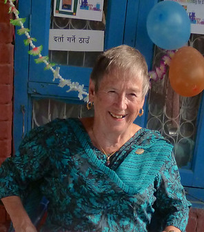 Elsie James smiling. She is in front of a door. Door has balloons and Nepali writing on it.