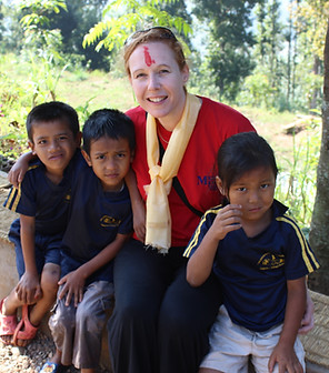 Karen Cuthbertson in Nepal. She is sitting with arms around 3 children. She is smiling, and has tika (red paste for ceremonies) on her forehead.