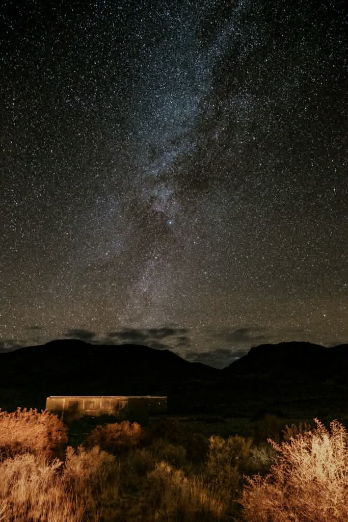 Starry night sky over mountains, with a glowing cabin and dry grass in the foreground. The Milky Way is prominently visible.