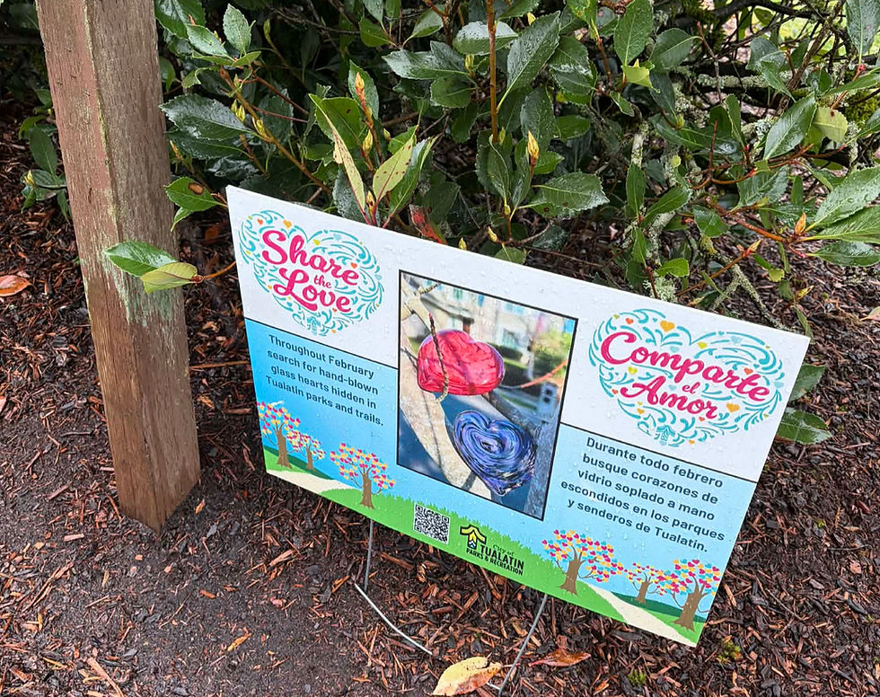 Sign in garden with colorful text "Share the Love" and "Comparte el Amor." It details a hunt for glass hearts in Tualatin parks.