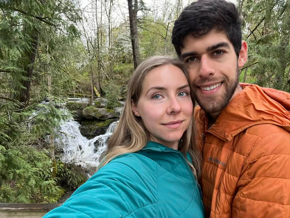 Couple in jackets smiles for a selfie in front of a forest waterfall. Green and orange clothing contrasts with lush greenery and flowing water.