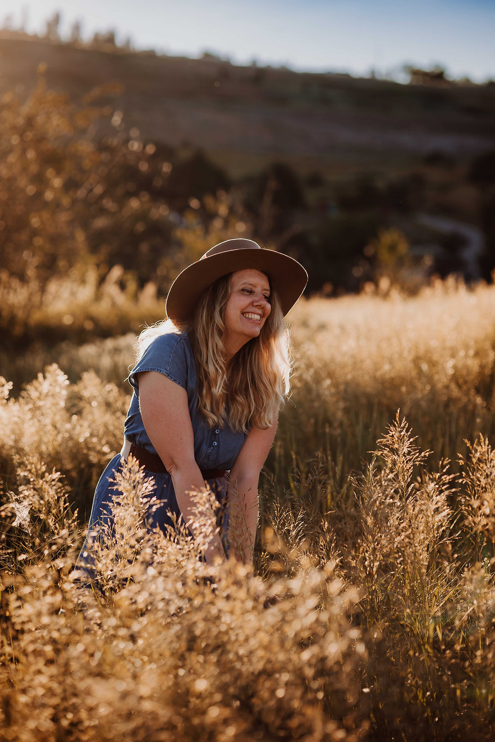 A smiling woman with long, wavy blonde hair and a wide-brimmed hat is standing in a golden field during sunset. She is wearing a denim dress and is looking to the side while gently bending down among the tall grass, creating a warm and joyful atmosphere.