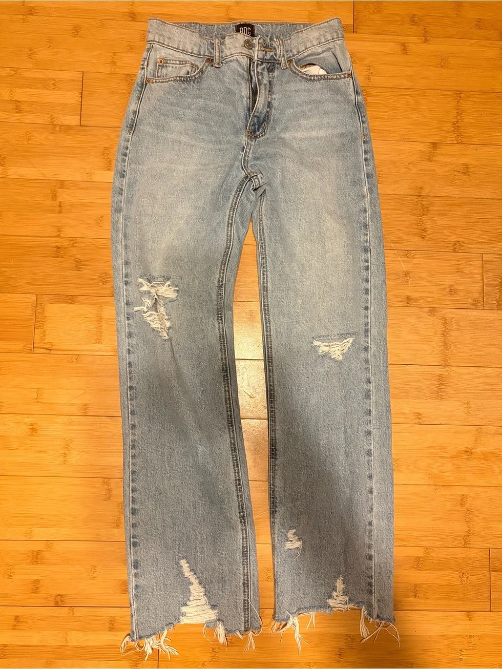 Light blue distressed jeans with frayed hems lie on a wooden floor, BNG label visible.