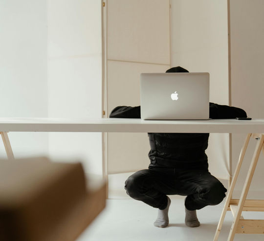 Person wearing dark clothing crouched behind a desk while using a laptop in a minimal modern workspace.