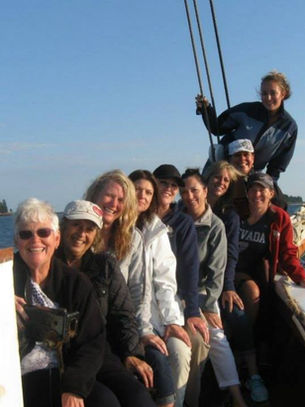 Group of women sitting together on the deck of the J. & E. Riggin schooner, smiling and bundled up while sailing along the Maine coast on an all-girls trip.
