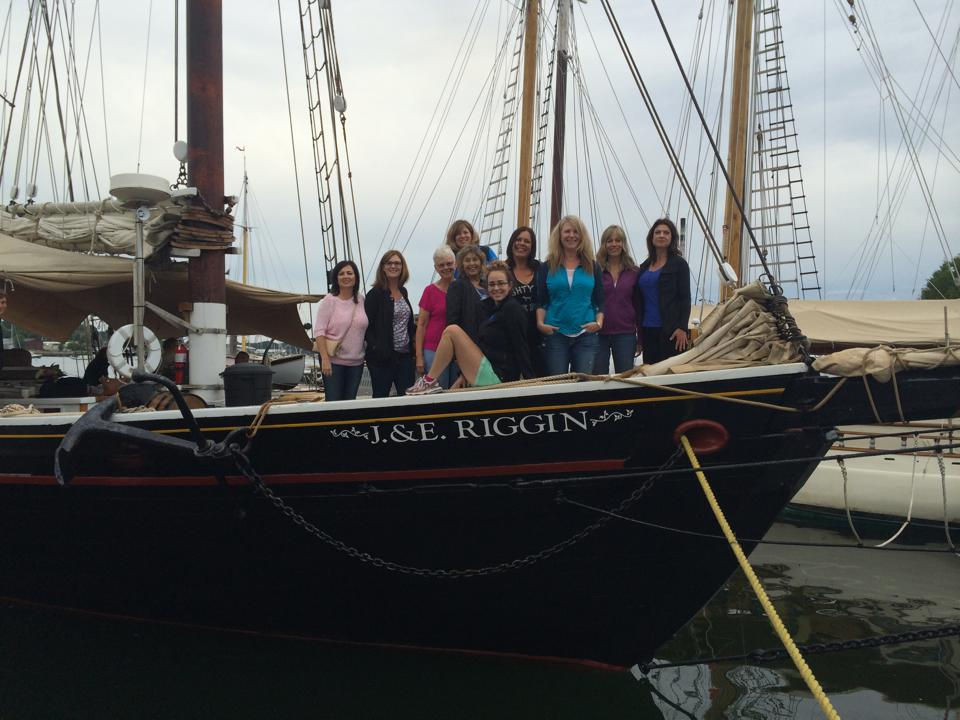 Group of women standing and sitting at the bow of the historic J. & E. Riggin schooner in harbor, ready for a Maine coast sailing adventure together.