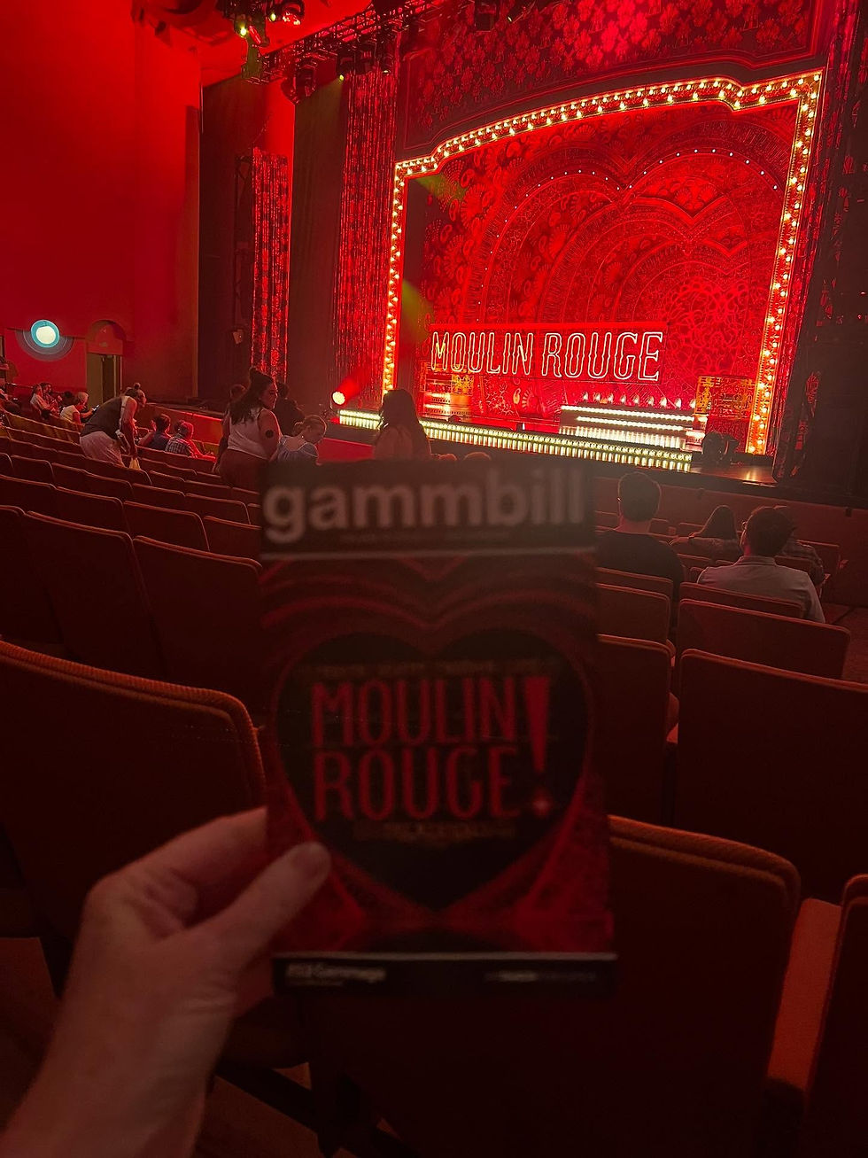View from the audience at ASU Gammage with a Moulin Rouge playbill in hand and the glowing red stage set in the background.